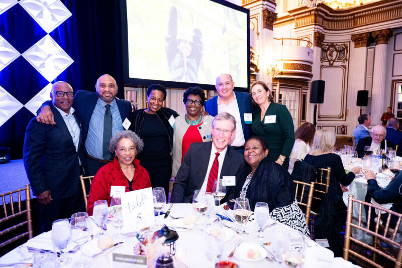 The image shows a group of people, likely at a formal event. They are gathered around a table, some smiling at the camera. There's a large screen in the background. The setting appears to be a banquet hall or similar venue. The people are dressed in formal attire.